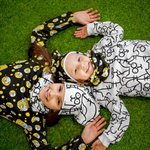 Dziewczynki ubrane w stroje Dzikie Pląsy kominy, czapki i bluzki, widok z góry, leżą na trawie, radosne miny, ubranka w koty w kolorach żółty, biały, czarny