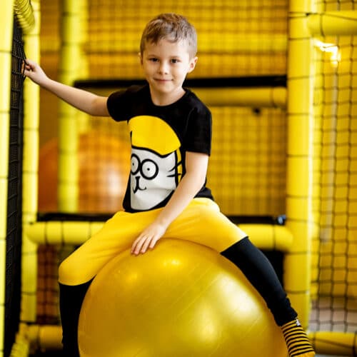 Chłopiec w stroju marki dzikei Pląsy na zjeżdżalni, żółte spodnie dresowe, czarna koszulka z kotem gigantem, skakanie na żółtej piłce